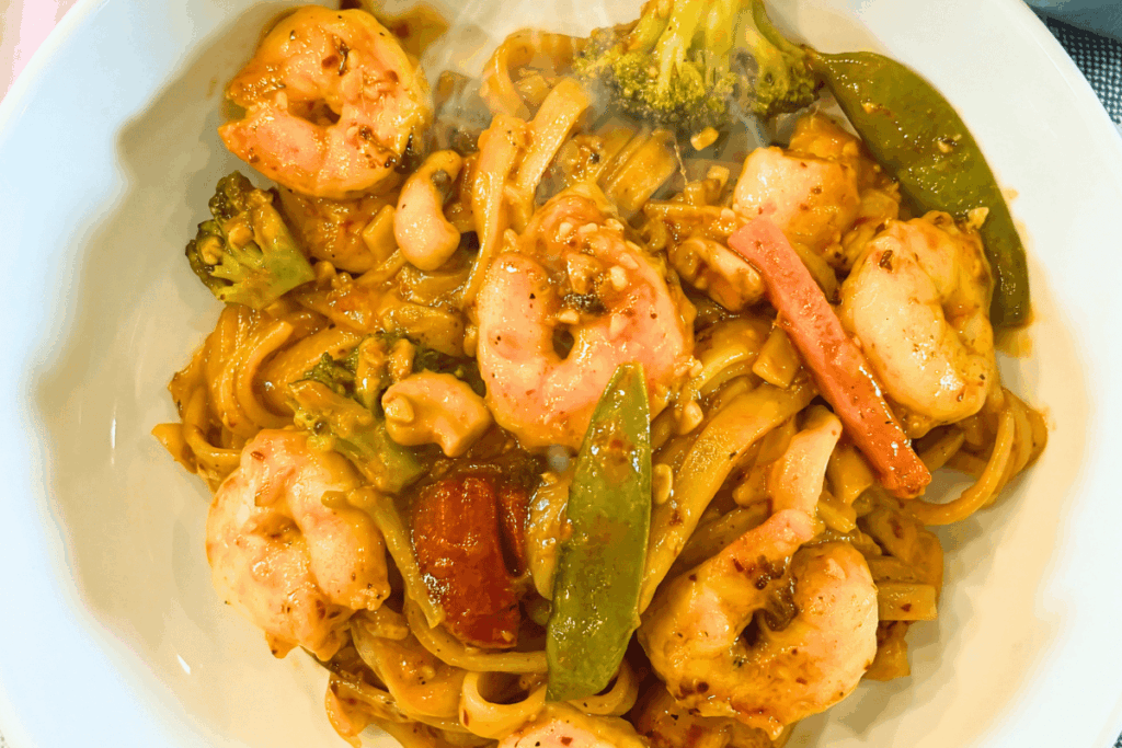 Top view of a white bowl with a serving of Chili Garlic Shrimp And Vegetable Noodles. Spicy, plump shrimp in a garlicky, chili sauce, with rice noodles, and Asian vegetables.